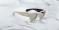 A pair of Jacques Marie Mage Chelsea beige, thick-framed sunglasses with dark lenses sits on a white marble surface with grey veins, with faint brand details visible on the inside of the left lens.