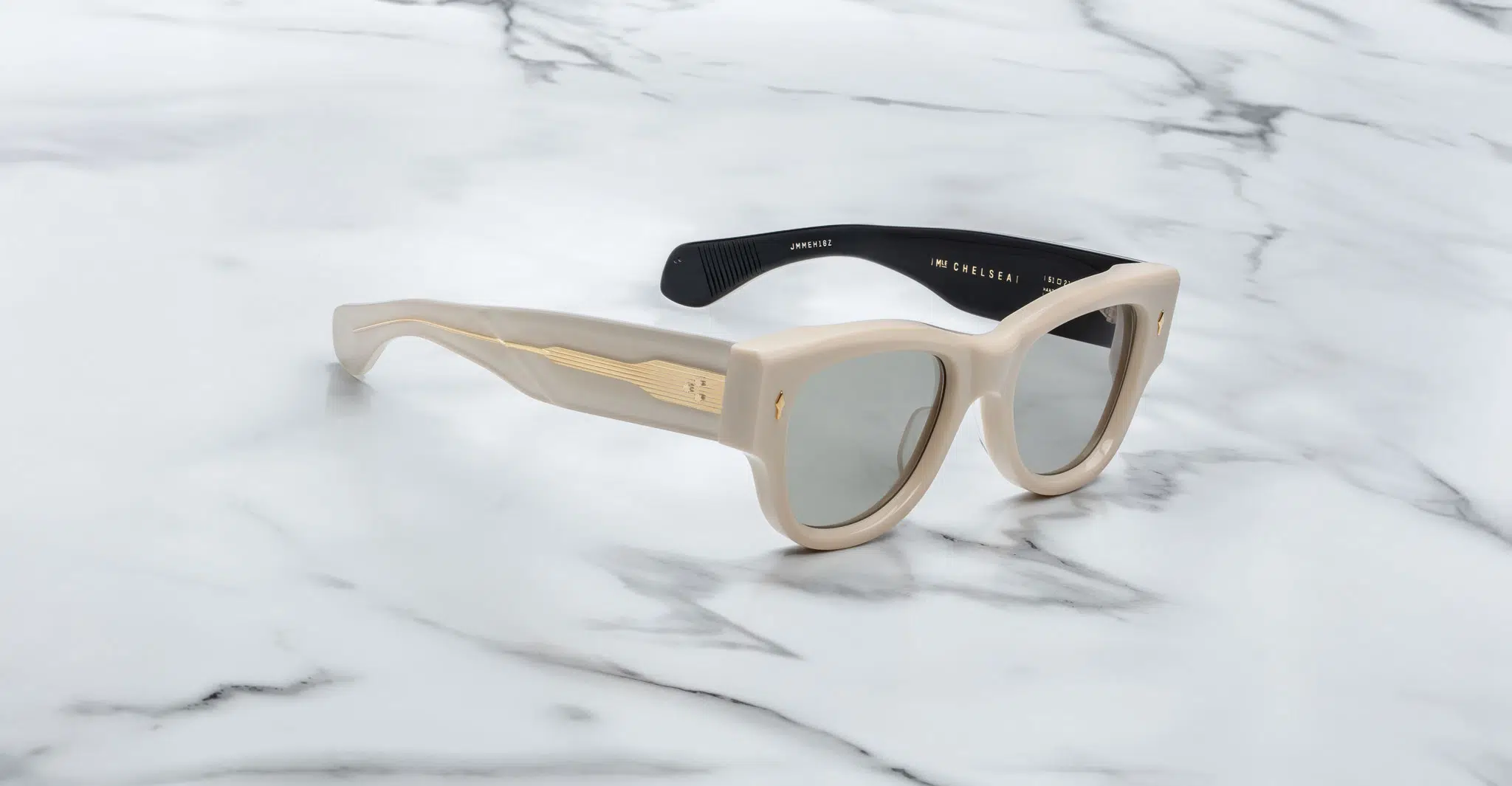 A pair of Jacques Marie Mage Chelsea beige, thick-framed sunglasses with dark lenses sits on a white marble surface with grey veins, with faint brand details visible on the inside of the left lens.