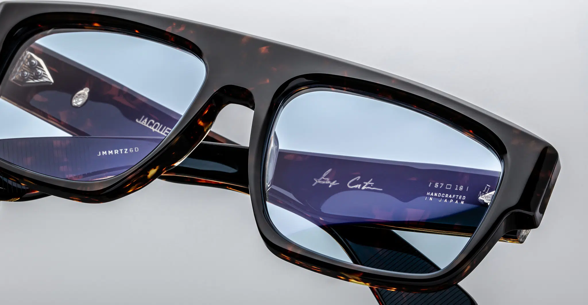 Close-up of Jacques Marie Mage Ritz tortoiseshell eyeglasses with blue-tinted lenses. Reflections reveal the interior frames, displaying white text with model numbers, brand name, and “handcrafted in Japan.”.