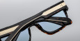 Close-up of two pairs of Jacques Marie Mage Ritz sunglasses in black with gold accents and blue-tinted lenses, stacked atop each other against a plain gray background.
