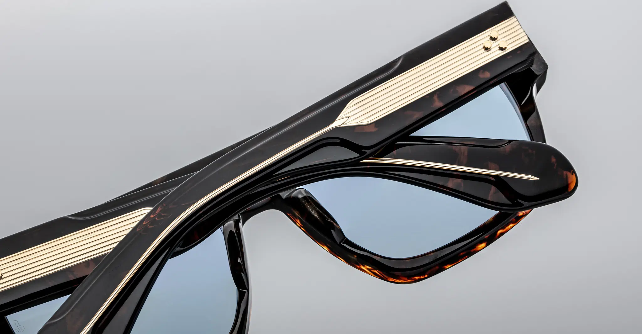 Close-up of two pairs of Jacques Marie Mage Ritz sunglasses in black with gold accents and blue-tinted lenses, stacked atop each other against a plain gray background.