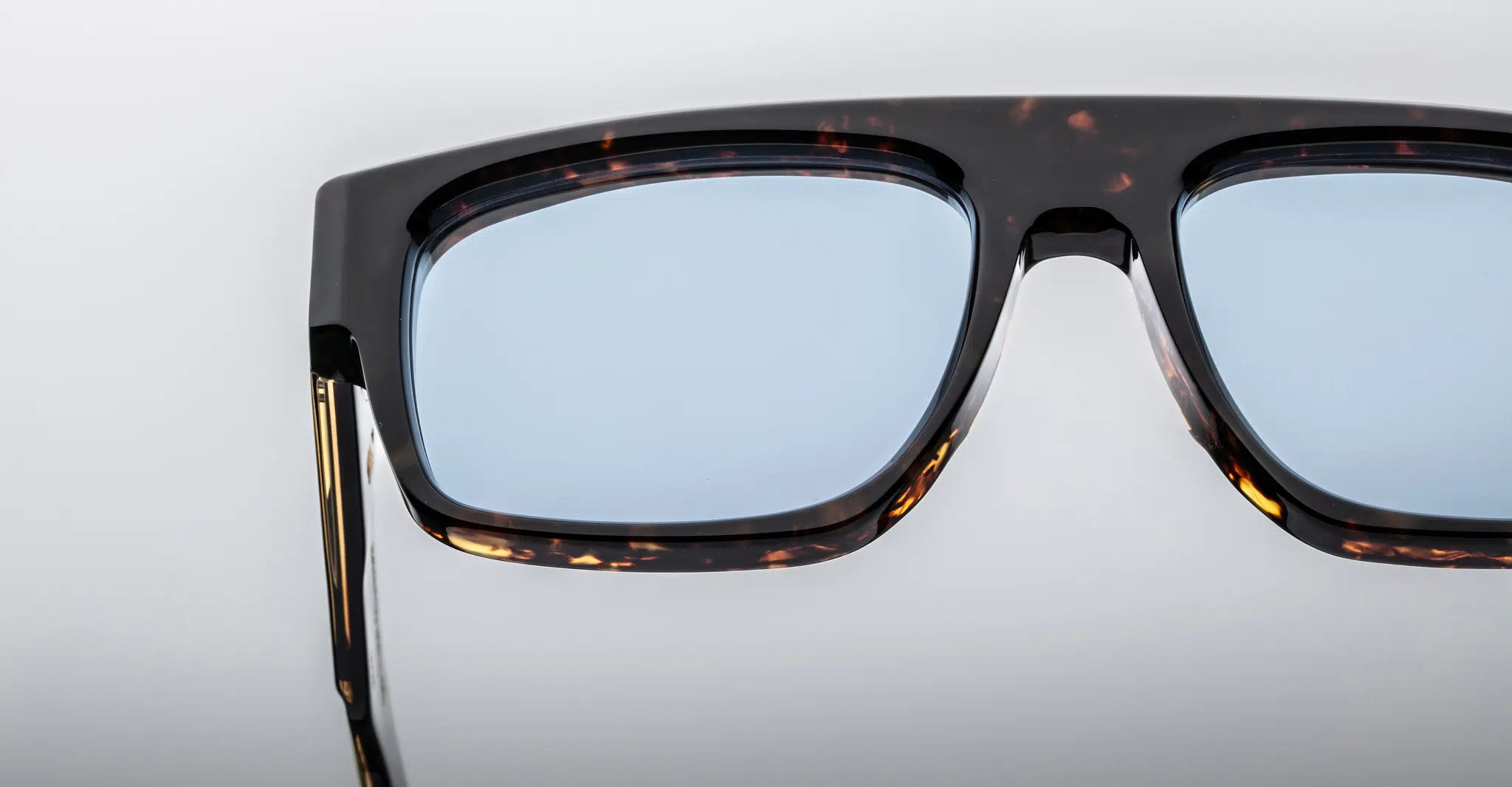 Close-up of the upper half of Jacques Marie Mage Ritz sunglasses, featuring a dark tortoiseshell frame and light blue lenses, set against a plain, light background.