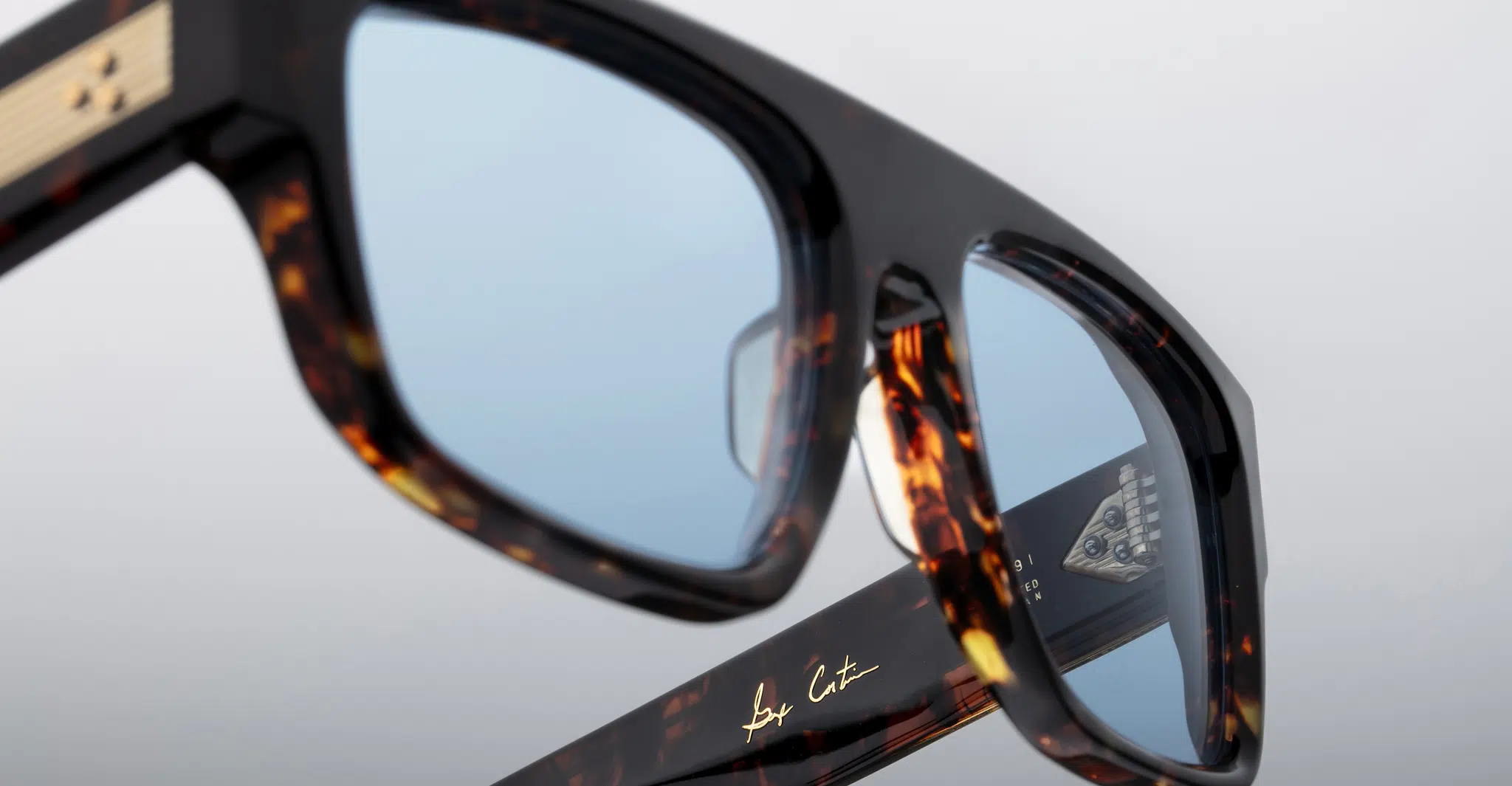 Close-up of Jacques Marie Mage Ritz tortoiseshell eyeglasses with blue-tinted lenses, gold detailing on the arms, and a signature reading by Cut on the inner side of one arm.