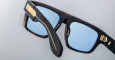 A close-up of Jacques Marie Mage Ritz sunglasses with black and brown tortoiseshell thick frames, blue-tinted lenses, and gold accents on the temple and near the hinge, set on a light gray surface.