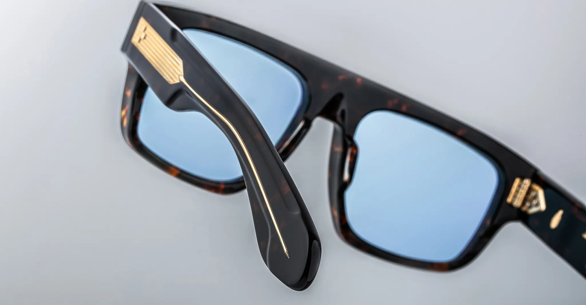 A close-up of Jacques Marie Mage Ritz sunglasses with black and brown tortoiseshell thick frames, blue-tinted lenses, and gold accents on the temple and near the hinge, set on a light gray surface.