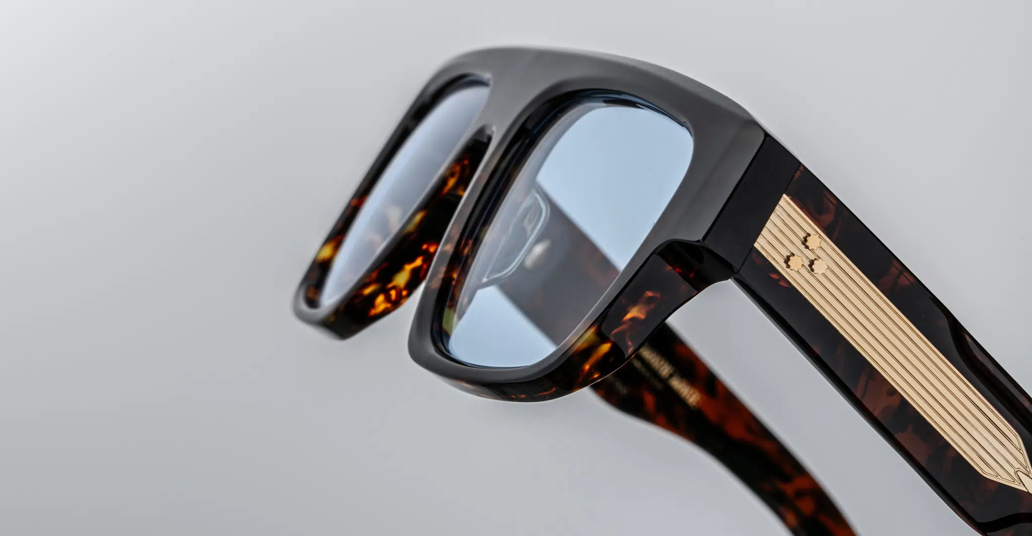 Close-up of Jacques Marie Mage Ritz eyeglasses featuring thick brown tortoiseshell frames, blue-tinted lenses, and gold detailing on the arms, set against a plain light gray background.