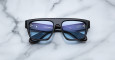 The Jacques Marie Mage Ritz sunglasses, featuring a black rectangular frame and blue-tinted lenses, rest face-up on a white marble surface with their inner arms and brand details clearly visible.