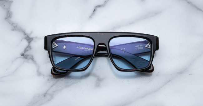 The Jacques Marie Mage Ritz sunglasses, featuring a black rectangular frame and blue-tinted lenses, rest face-up on a white marble surface with their inner arms and brand details clearly visible.