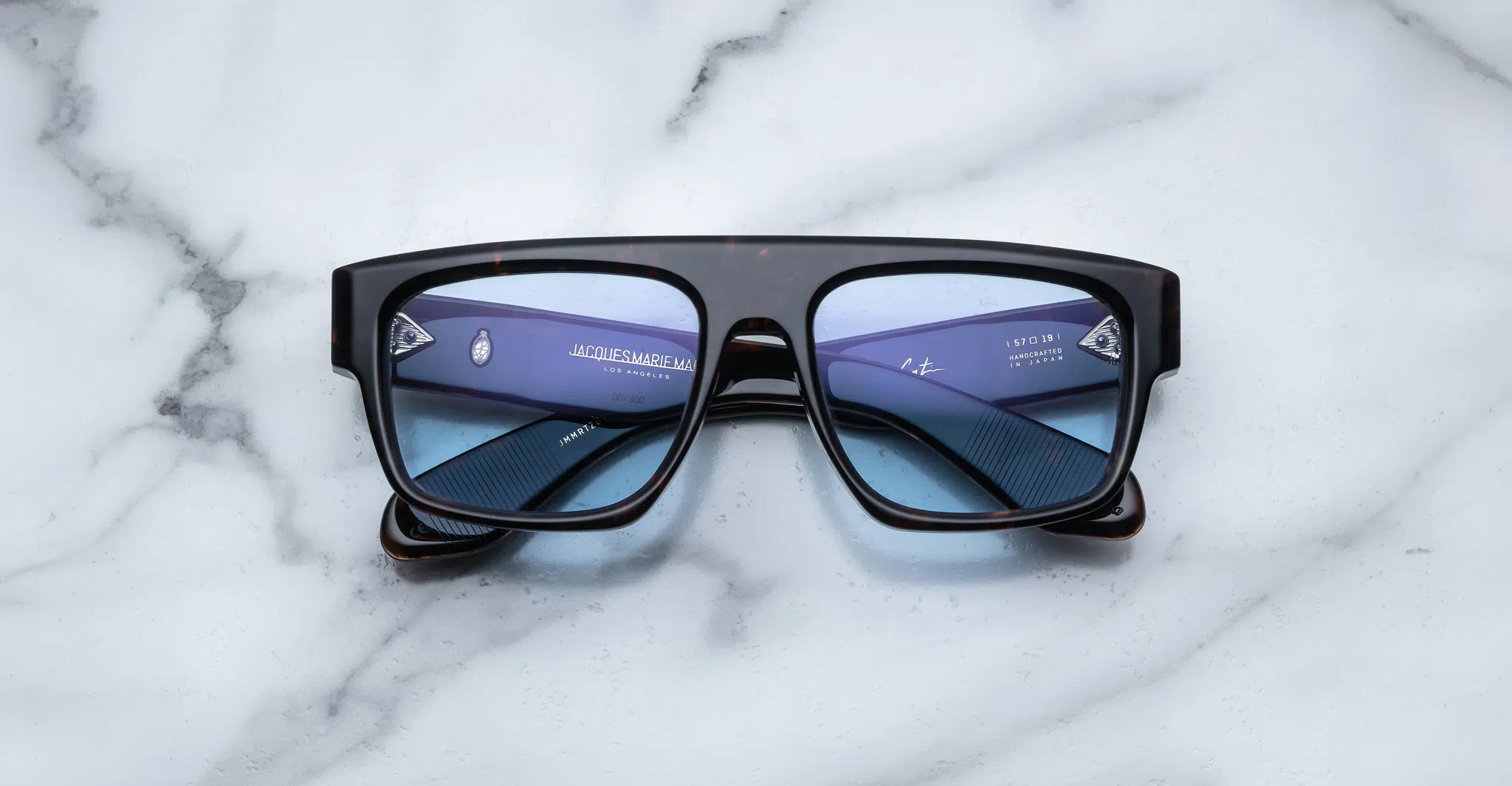 The Jacques Marie Mage Ritz sunglasses, featuring a black rectangular frame and blue-tinted lenses, rest face-up on a white marble surface with their inner arms and brand details clearly visible.