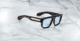 The Jacques Marie Mage Ritz are thick tortoiseshell sunglasses with blue-tinted lenses and gold accents on the temples, displayed on a white marble surface.