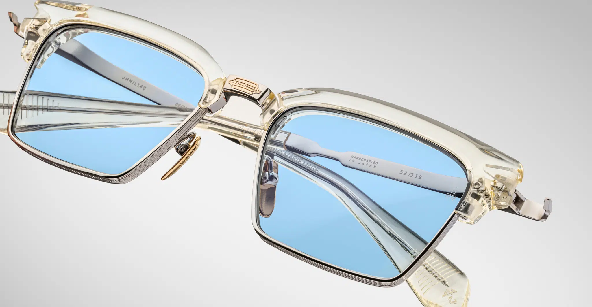 Close-up of Jacques Marie Mage Alin: stylish, translucent square eyeglasses with light blue lenses and gold accents, set against a plain background. The modern frames have a sleek, refined look and high-quality finish.