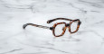 Jacques Marie Mage Holden eyeglasses featuring tortoiseshell-patterned, geometric, slightly oversized frames with metallic hinges, displayed on a white marble surface.