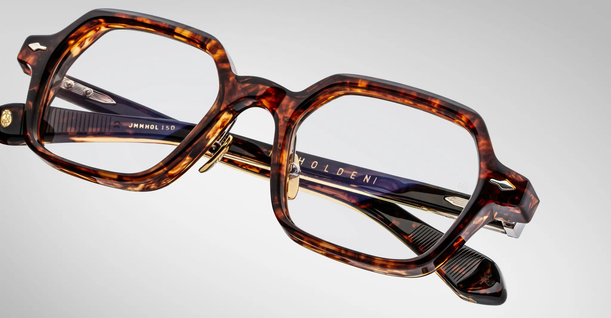 Jacques-Marie-Mage-Holden-Samoa-JMMHOLRX-15D-Detail1-Dan-Deutsch Close-up of Jacques Marie Mage Holden rectangular tortoiseshell eyeglasses with thick frames and gold accents, showing the brand name on the inner arm, set against a plain light background.