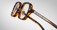 Close-up of Jacques Marie Mage Holden tortoiseshell eyeglasses featuring thick square frames and metallic hinges, set against a plain light gray background.