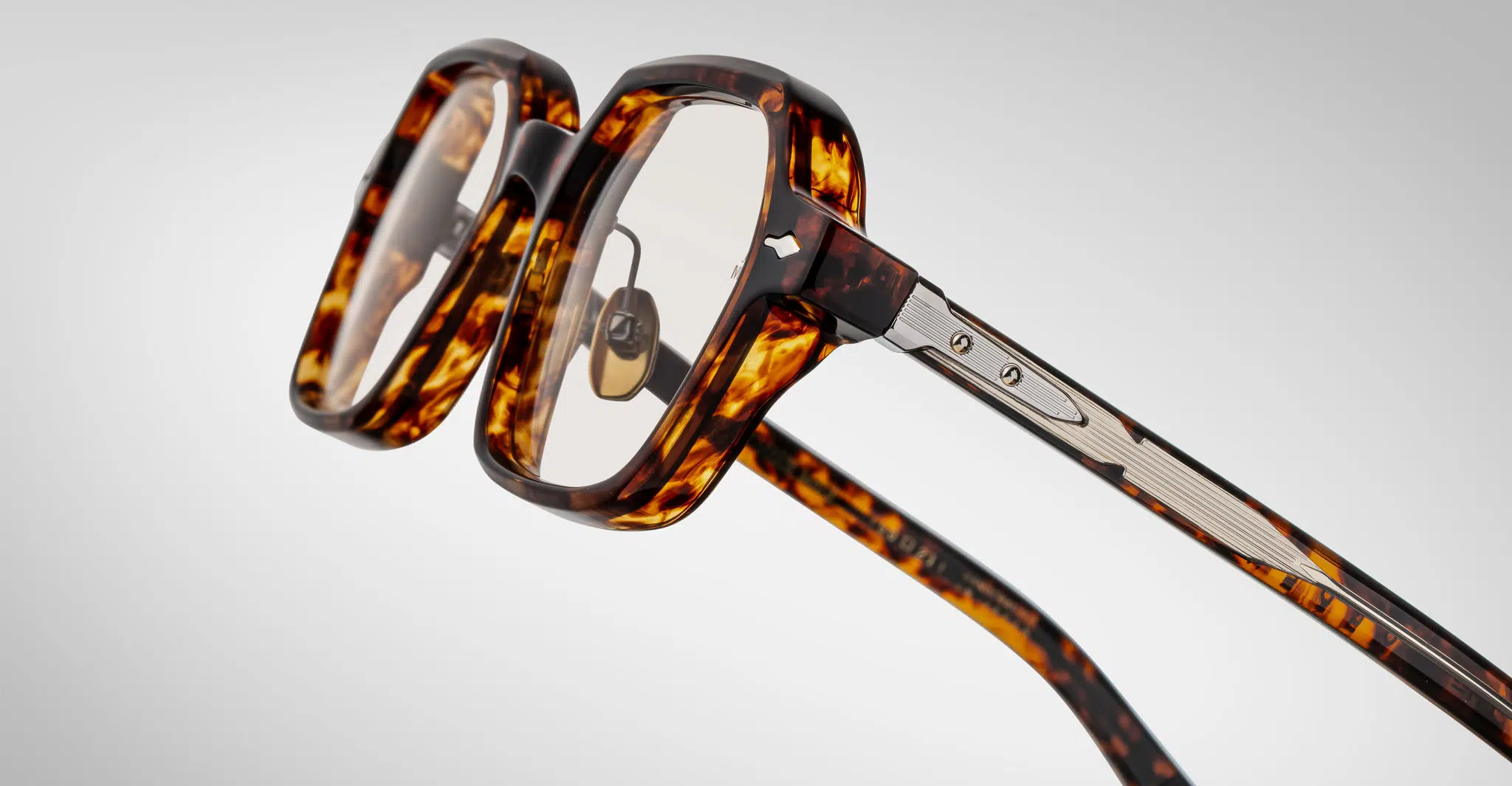 Jacques-Marie-Mage-Holden-Samoa-JMMHOLRX-15D-Detail2-Dan-Deutsch Close-up of Jacques Marie Mage Holden tortoiseshell eyeglasses featuring thick square frames and metallic hinges, set against a plain light gray background.