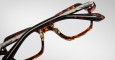 A close-up of two Jacques Marie Mage Holden eyeglasses with tortoiseshell frames and metallic hinges, stacked diagonally on a plain light gray background.