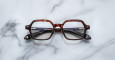 The Jacques Marie Mage Holden eyeglasses, featuring brown tortoiseshell square frames and black arms, rest on a white marble surface with gray veins.