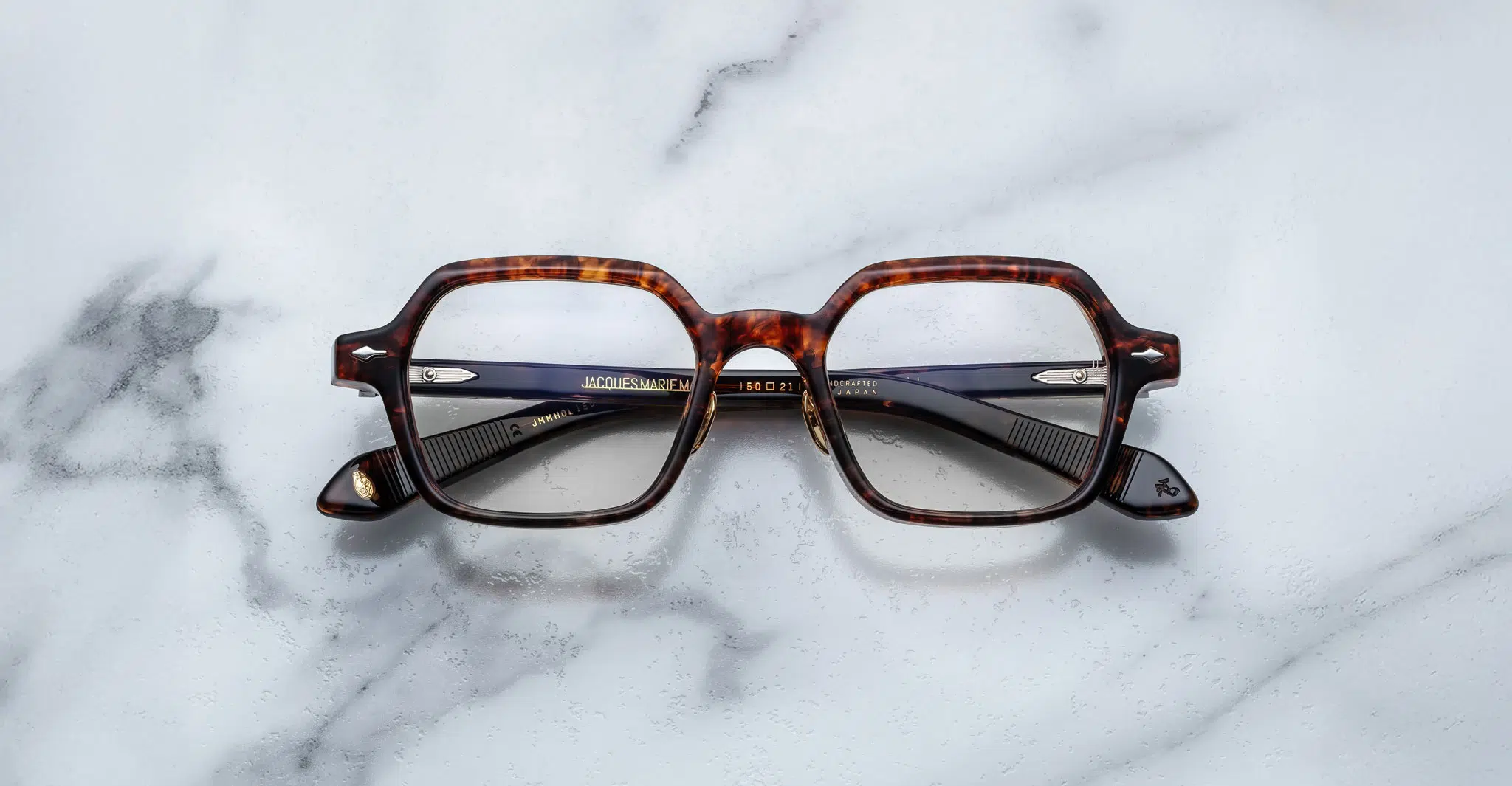 Jacques-Marie-Mage-Holden-Samoa-JMMHOLRX-15D-Front-Dan-Deutsch The Jacques Marie Mage Holden eyeglasses, featuring brown tortoiseshell square frames and black arms, rest on a white marble surface with gray veins.