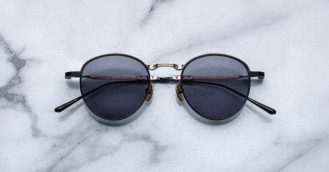 A pair of round, moon gold-framed sunglasses with charcoal lenses is placed on a white marble surface. The arms of the sunglasses are metallic with visible branding on the inner side of the arms.