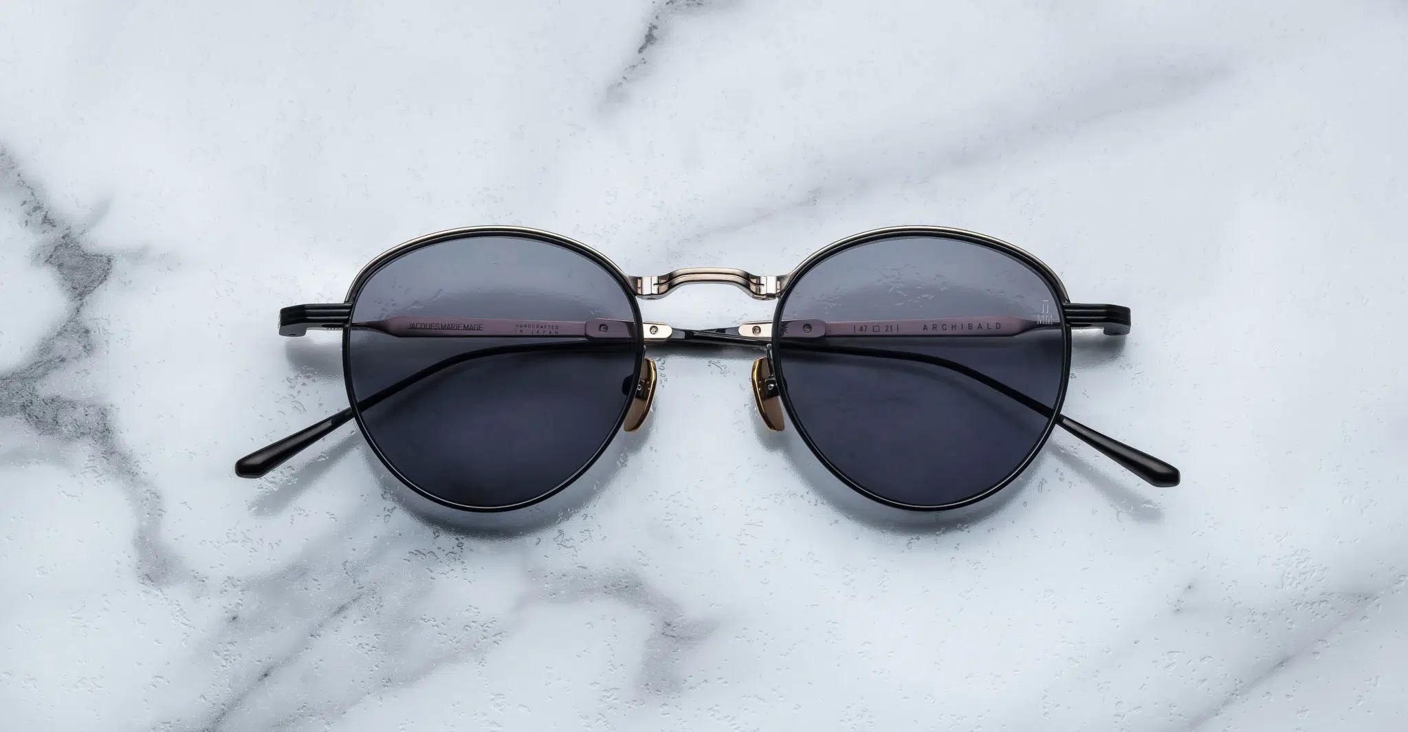 A pair of round, moon gold-framed sunglasses with charcoal lenses is placed on a white marble surface. The arms of the sunglasses are metallic with visible branding on the inner side of the arms.
