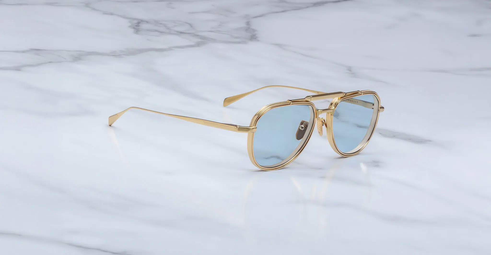 The Jacques Marie Mage Commander gold-framed aviator sunglasses with light blue lenses rest on a white marble surface adorned with gray veining.