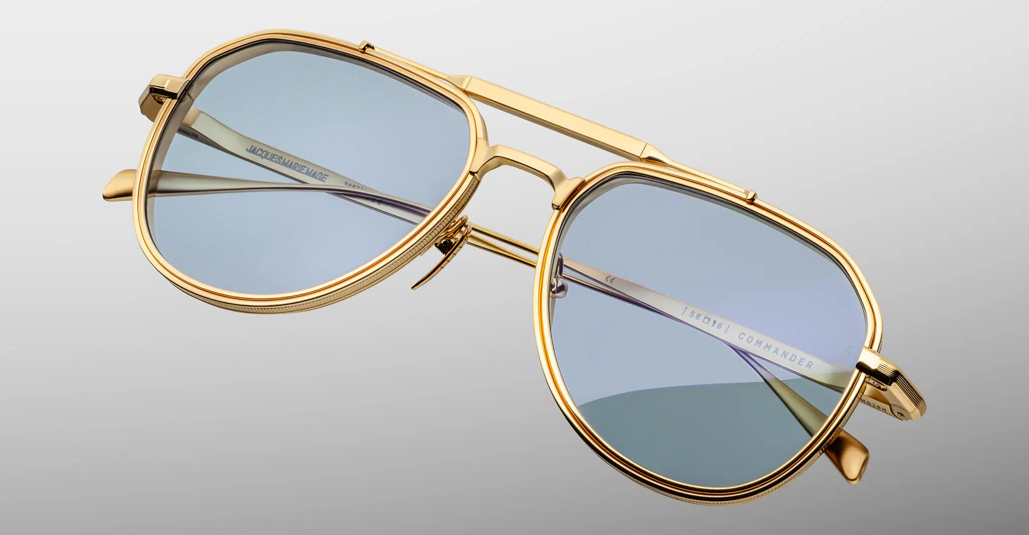 A pair of gold-framed aviator sunglasses with light blue tinted lenses, named Jacques Marie Mage Commander, is shown against a light gray background. The brand name is visible on the inside of the arm.