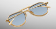 The Jacques Marie Mage Commander sunglasses, featuring gold frames and blue-tinted lenses, are displayed upside down with folded arms against a smooth gradient background from light gray to white.