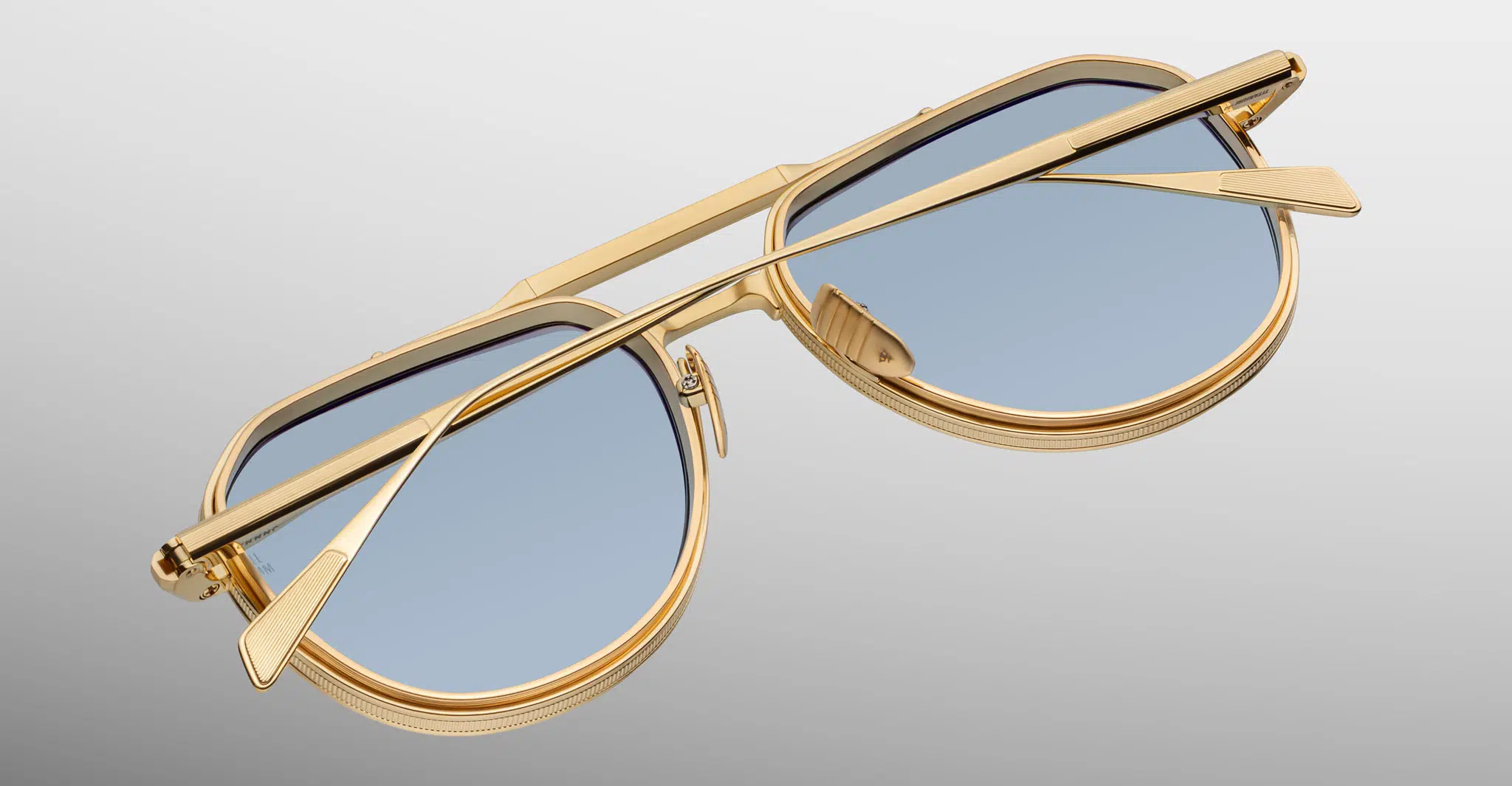The Jacques Marie Mage Commander sunglasses, featuring gold frames and blue-tinted lenses, are displayed upside down with folded arms against a smooth gradient background from light gray to white.