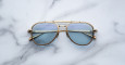 A pair of Jacques Marie Mage Commander sunglasses with gold frames and light blue lenses rests on a white marble surface, reflecting the marble pattern in the lenses.