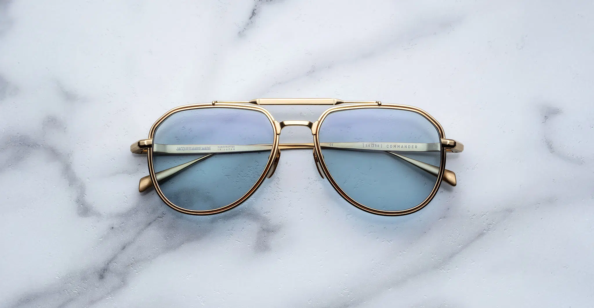 A pair of Jacques Marie Mage Commander sunglasses with gold frames and light blue lenses rests on a white marble surface, reflecting the marble pattern in the lenses.