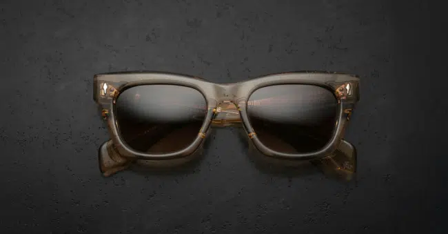 The Jacques Marie Mage Dealan sunglasses, featuring brown translucent square frames and dark lenses, are displayed from the front on a textured dark surface.