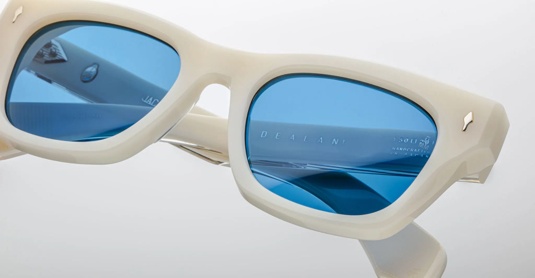 Close-up of cream-colored Jacques Marie Mage Dealan sunglasses with blue tinted lenses, metallic accents on the frame and temple corners, and “DEALAN” visible on the inside of one arm.