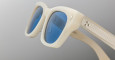 Close-up of Jacques Marie Mage Dealan sunglasses with thick off-white frames, blue-tinted lenses, metallic-accented temples, and a grooved design, set against a neutral gradient background.