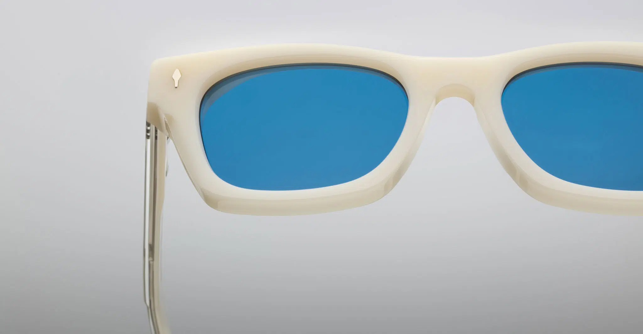 Close-up of the top half of Jacques Marie Mage Dealan white rectangular sunglasses with blue-tinted lenses on a plain light gray background. A small gold emblem appears near the temple on the left side of the frame.