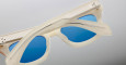 Close-up of Jacques Marie Mage Dealan sunglasses in beige with translucent frames and blue lenses, shown folded with arms crossed on a plain light gradient background.