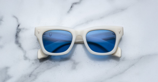 The Jacques Marie Mage Dealan sunglasses, featuring thick white rectangular frames and blue-tinted lenses, rest on a white marble surface with gray veins.