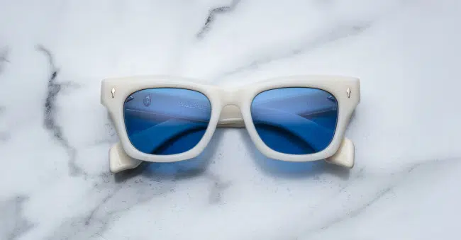 The Jacques Marie Mage Dealan sunglasses, featuring thick white rectangular frames and blue-tinted lenses, rest on a white marble surface with gray veins.