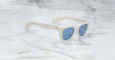 The Jacques Marie Mage Dealan sunglasses, featuring white frames and blue-tinted lenses, are angled on a white marble surface with grey veins, showcasing the left arm and front of the stylish frame.