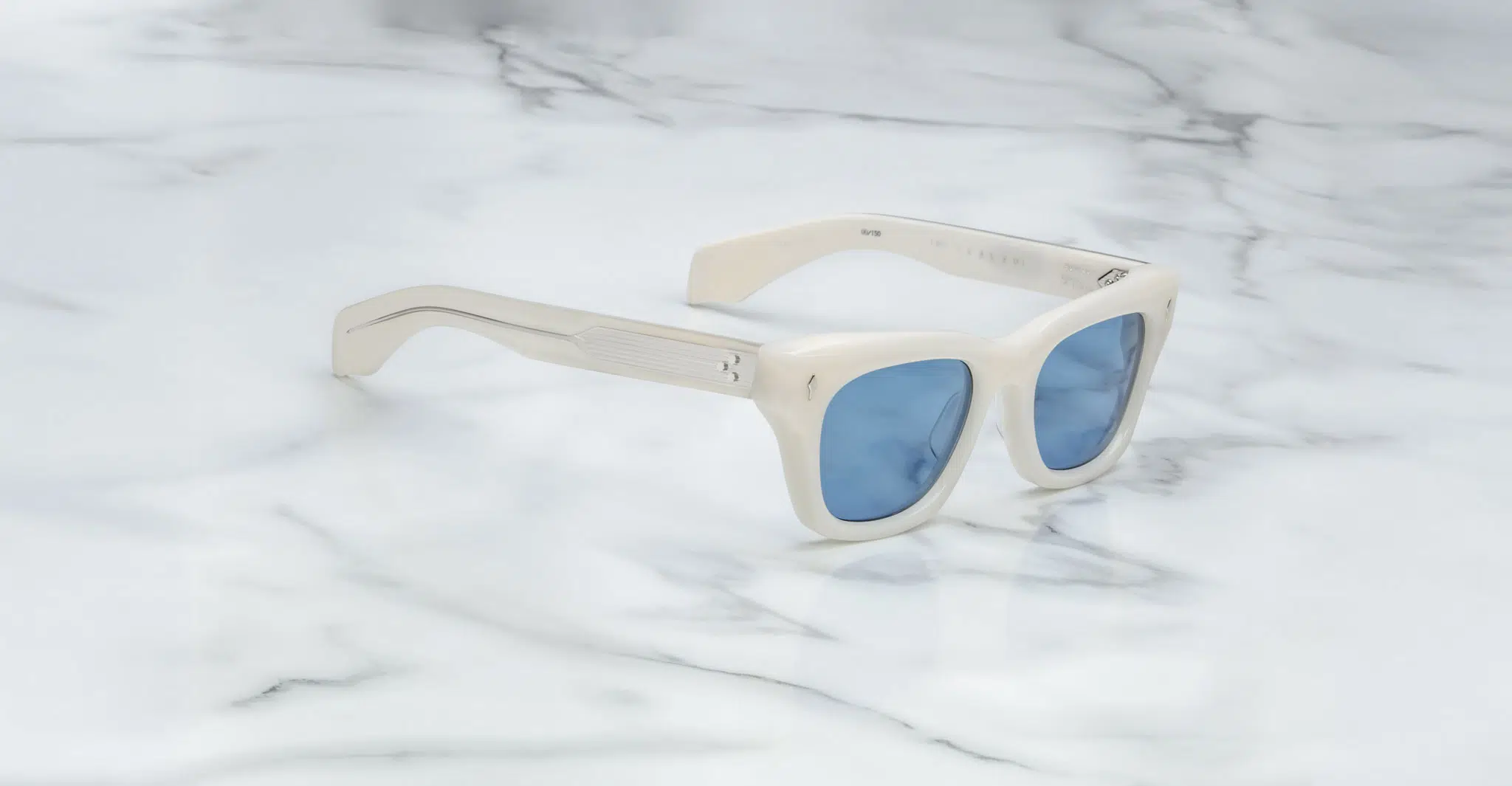 The Jacques Marie Mage Dealan sunglasses, featuring white frames and blue-tinted lenses, are angled on a white marble surface with grey veins, showcasing the left arm and front of the stylish frame.