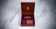 A hinged wooden box with an ornate emblem inside the lid, reminiscent of Jacques Marie Mage Decades Packaging, opens to reveal a screwdriver, rectangular card, and envelope in marble-set compartments.