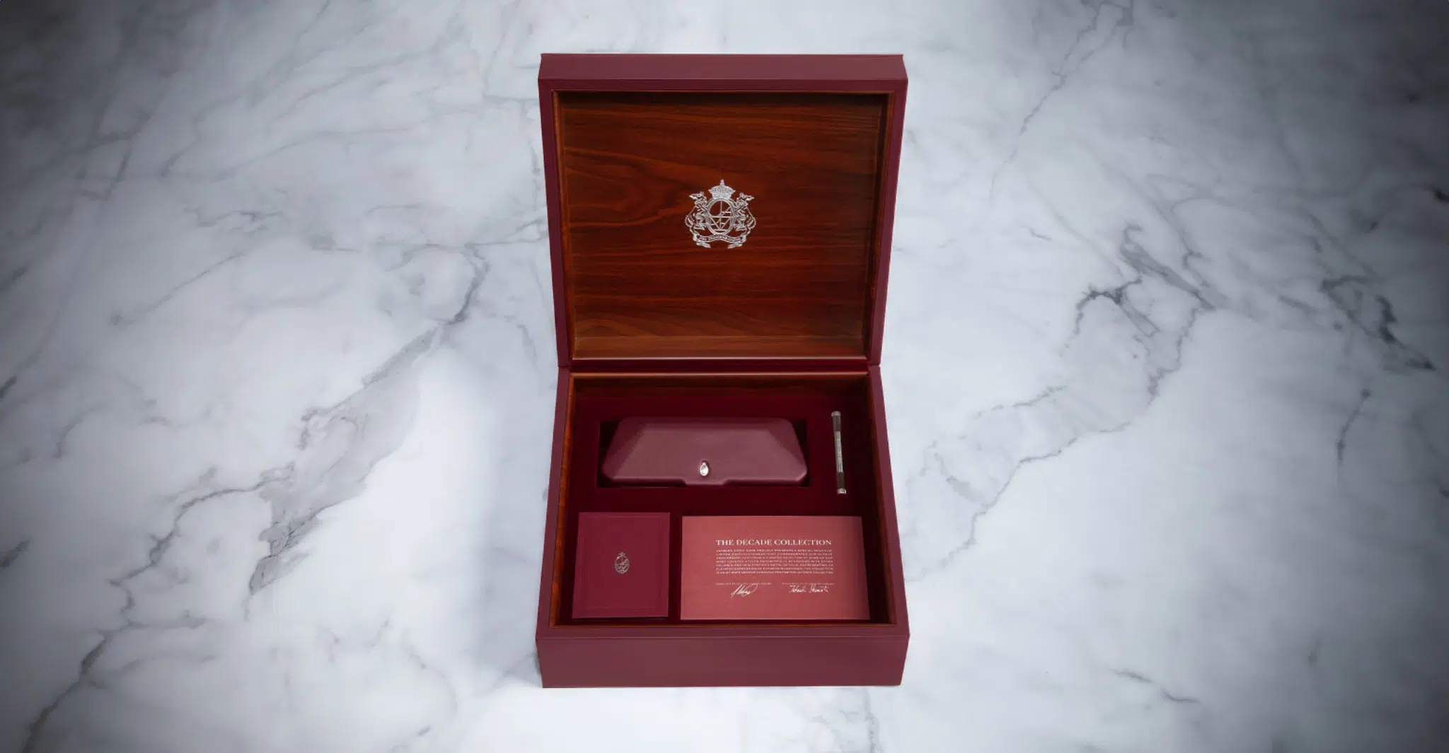 A hinged wooden box with an ornate emblem inside the lid, reminiscent of Jacques Marie Mage Decades Packaging, opens to reveal a screwdriver, rectangular card, and envelope in marble-set compartments.