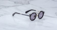 The Jacques Marie Mage Delage sunglasses, featuring round black frames with silver accents and dark lenses, are displayed on a white marble surface.