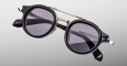 The Jacques Marie Mage Delage sunglasses feature round black frames with dark tinted lenses, silver accents, and a double bridge, displayed on a light gray background. The brand name is visible on the black arms.