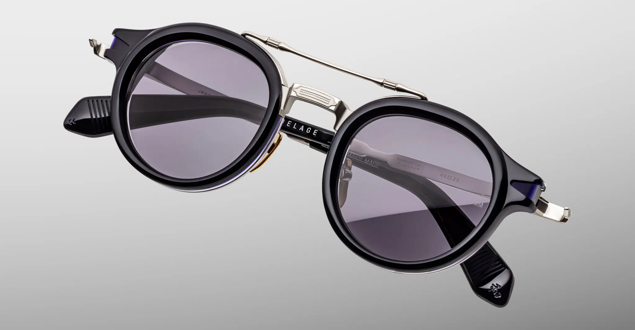 The Jacques Marie Mage Delage sunglasses feature round black frames with dark tinted lenses, silver accents, and a double bridge, displayed on a light gray background. The brand name is visible on the black arms.