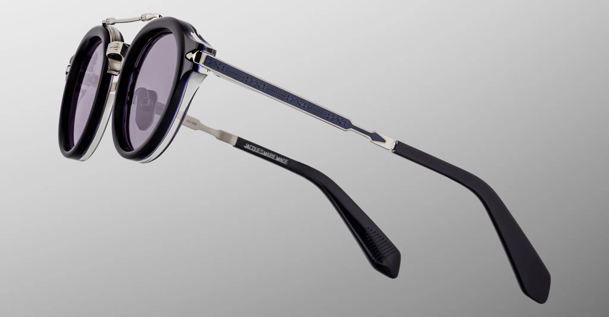 A close-up side view of Jacques Marie Mage Delage sunglasses featuring round dark lenses, thin blue arms with silver accents, and the brand name visible on the arm, set against a gradient gray background.