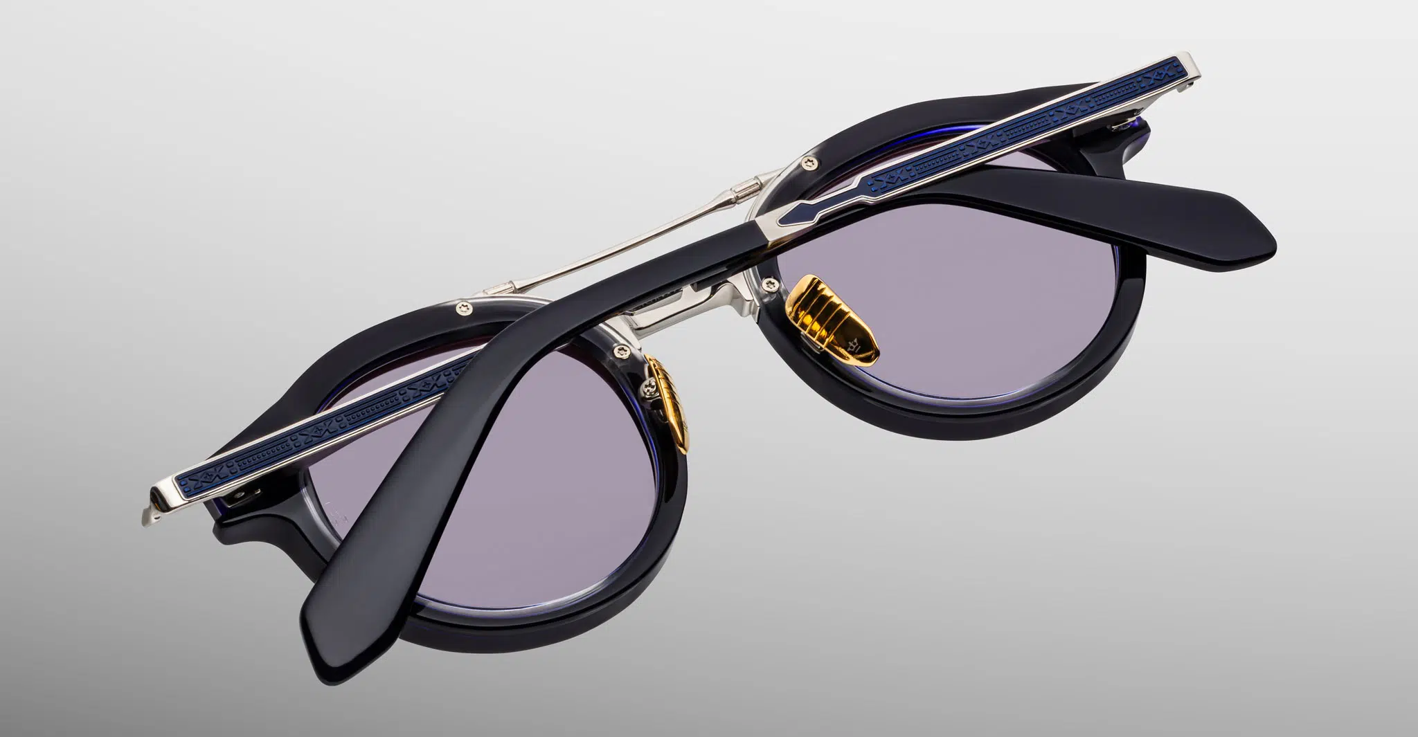 The Jacques Marie Mage Delage sunglasses are shown folded with crossed temples, featuring round black frames, purple-tinted lenses, and gold nose pads against a plain light gradient background.