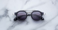 The Jacques Marie Mage Delage, a pair of round black sunglasses with dark lenses and silver accents, rests on a white marble surface featuring subtle grey veining.