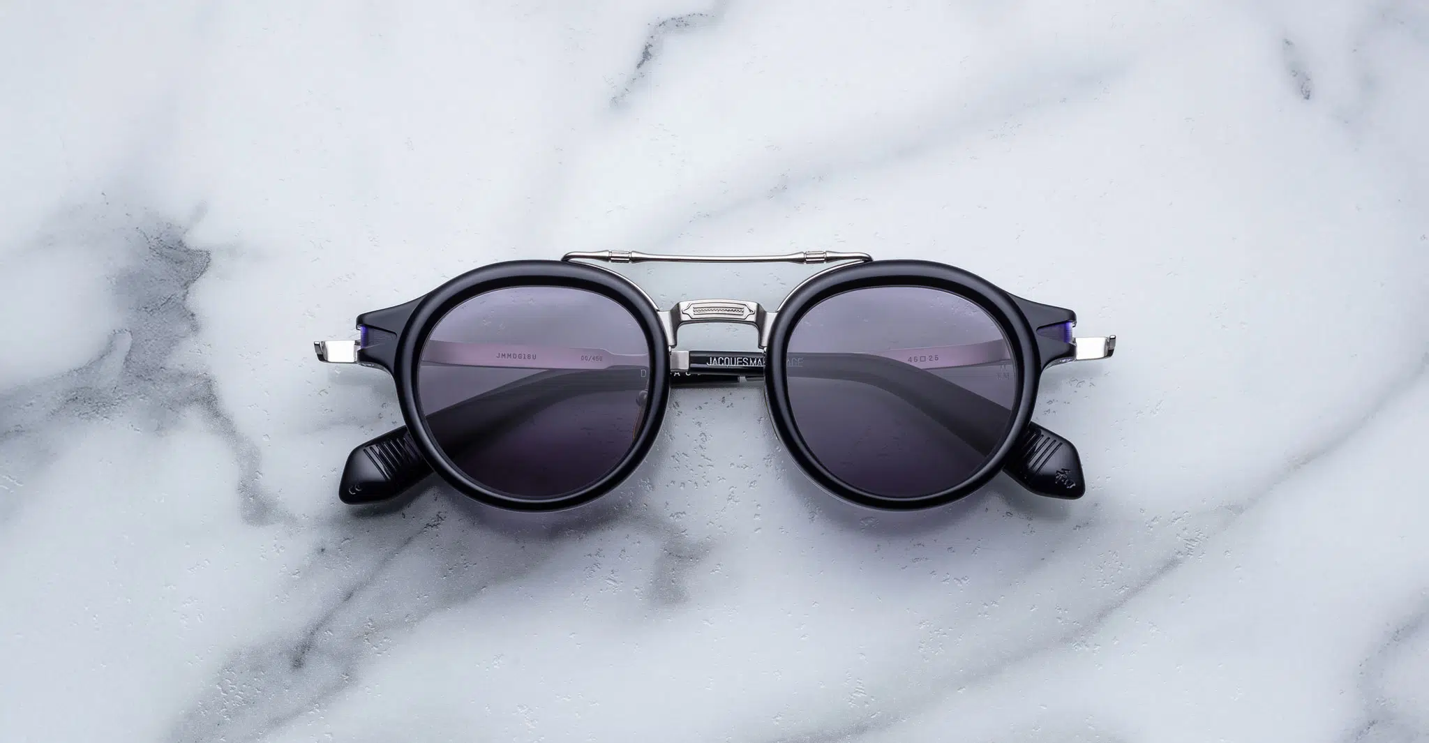 The Jacques Marie Mage Delage, a pair of round black sunglasses with dark lenses and silver accents, rests on a white marble surface featuring subtle grey veining.