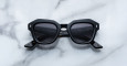 A pair of Jacques Marie Mage Eichler black, thick-framed sunglasses with grey lenses rests on a white marble surface with grey veins, with faint brand details visible on the inside of the left lens.