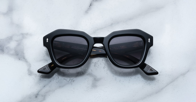A pair of Jacques Marie Mage Eichler black, thick-framed sunglasses with grey lenses rests on a white marble surface with grey veins, with faint brand details visible on the inside of the left lens.